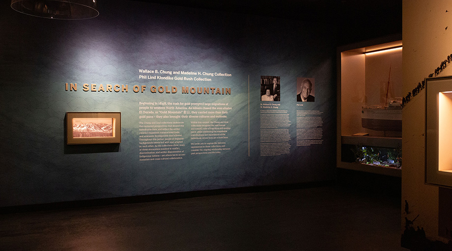 A photo of the gallery entrance, with text on the wall reading "In Search of Gold Mountain" and photos of Wallace and Madeline Chung and Phil Lind.
