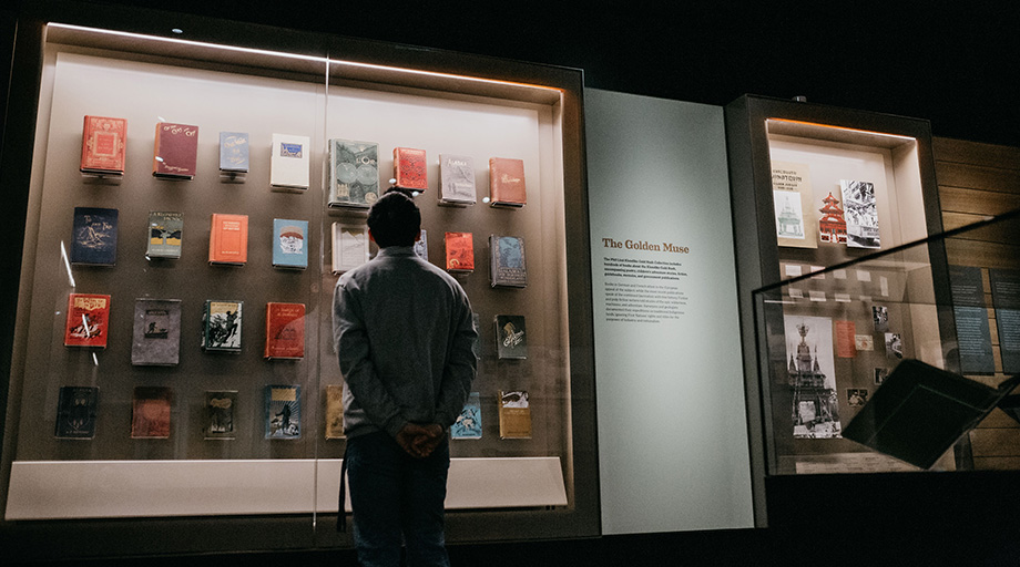 A photo of a gallery visitor looking at a display of books mounted on wall behind glass.